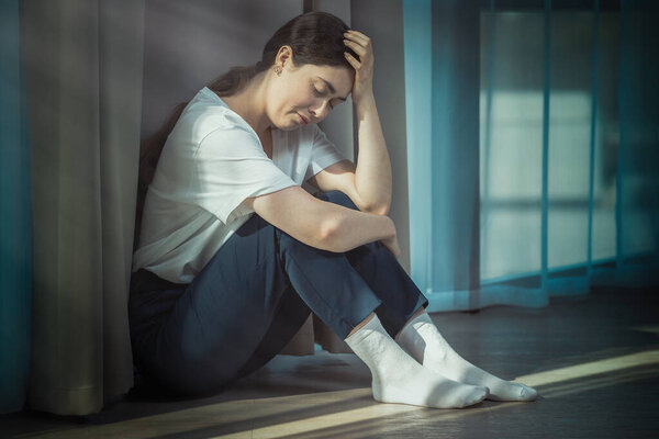 Alone depressed woman holding her head with her hand and sitting on the floor. Dark colors. The concept of domestic violence.