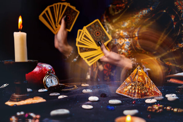 Астрология и эзотерика. Волшебная стеклянная пирамида с волшебным свечением. На заднем плане гадалка держит фаната карт Таро. Темный стол с рунами и свечами. Крупный план.