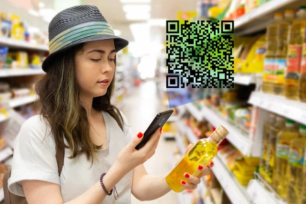 Young beautiful woman chooses a bottle of oil in the supermarket and checks the QR code on the label. The concept of modern technology and shopping in the store.