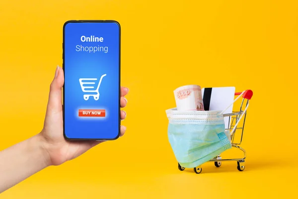 A shopping cart with a medical mask on, filled with cash and a Bank card. A woman\'s hand holds a smartphone. Yellow background. Virus protection and online purchase of medicines.