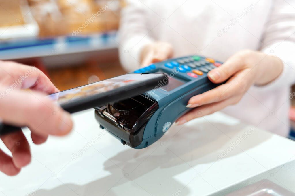 A woman's hands are held by a payment terminal and a man pays for a purchase using a smartphone. The concept of NFC, business and banking transactions.