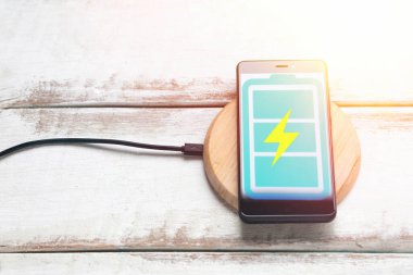 Modern teknoloji kavramı. Kablosuz şarjda turkuaz pil ikonlu siyah bir akıllı telefon var. Yatay çizgili beyaz ahşap arka plan. Uzayı kopyala Düz çizgi. Işık.