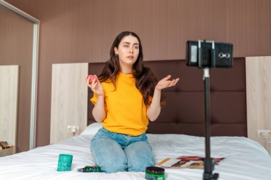 Vloglama. Genç ve güzel bir kadın vlogger akıllı telefondan canlı yayın yapıyor yatakta oturuyor ve kozmetik hakkında konuşuyor..