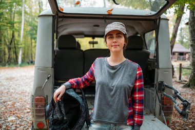 Beyaz bir kadın sırt çantasıyla bir cipin bagajına poz veriyor. Arka planda ağaçlar ve orman var. Kapatın. Araba yolculuğu ve turizm kavramı.