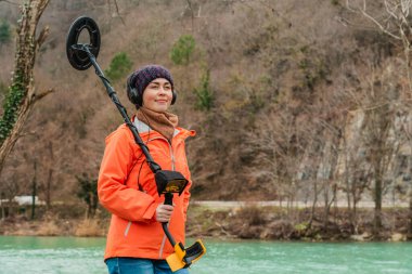 Search With A Metal Detector. A young woman posing with a metal detector on the background of the river. Close up. clipart