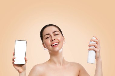 Kafkasyalı güzel bir kadının portresi elinde akıllı bir telefonla termal ferahlatıcı su püskürtüyor. Bej arka plan. Boşluğu kopyala. Güzellik, teknoloji ve kendine bakım kavramı..