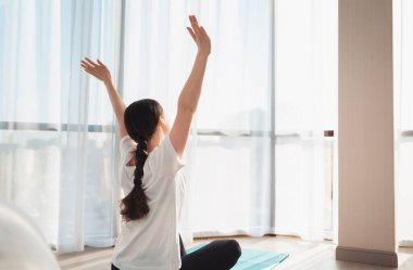 Yoga. Genç bir kadın spor minderinde oturuyor, pencereden dışarı bakıyor ve sabah güneşinin tadını çıkarıyor. Arka görünüm.