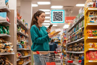 Beyaz bir kadın akıllı telefonuyla yiyecek paketinin QR kodunu tarıyor. Ürünün üstündeki QR kod simgesi. Arka planda bir süpermarket var. Modern teknoloji ve alışveriş kavramı.