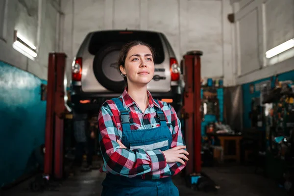 制服を着た若い女性の笑顔の整備士の肖像画は 彼女の腕でポーズを交差した 背景には自動車修理工場があります — ストック写真