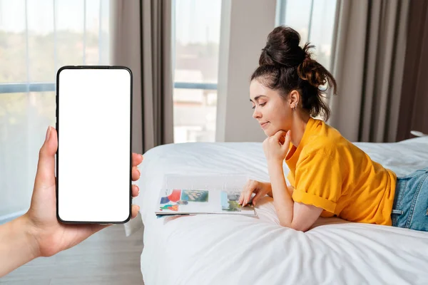 Online advertising. A young smiling woman is lying on the bed and reading a magazine. Home environment. Hand with smartphone. Mock up. Side view.