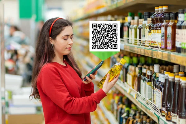 Beyaz bir kadın akıllı telefonuyla bir yağ şişesinin QR kodunu tarıyor. Ürünün üstündeki QR kod simgesi. Arka planda bir süpermarket var. Modern teknoloji ve alışveriş kavramı.
