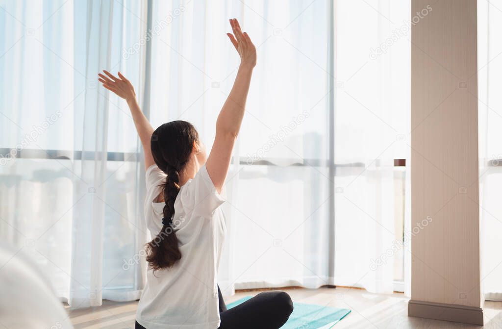 Yoga. A young woman is sitting on a sports mat, looking out the window and raising her hands up, enjoying the morning sun. Rear view.