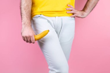 Potency and men's health. A man in white jeans is holding a banana near his genitals with his legs together and his hand on his waist. Pink background. Close up. Copy space. clipart