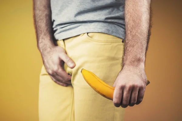 Impotency and masculine strength. A man in yellow jeans, holding a banana in his hand, at the level of the genitals, covering the genitals with his hand. Dark yellow background. Close up.