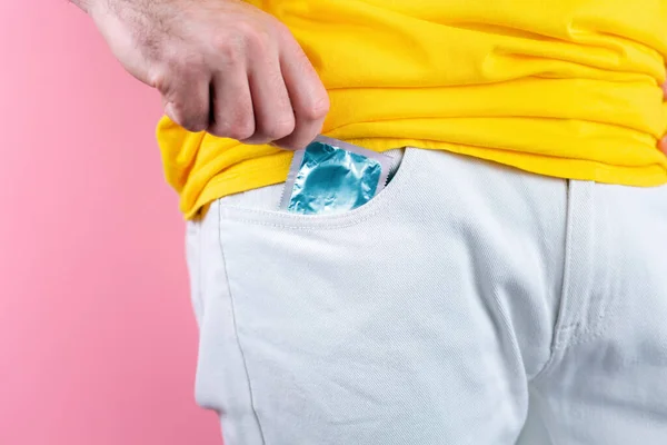 A man in white jeans pulls a condom out of his pocket. Close up. Pink background. Concept of protection against sexually transmitted infections.