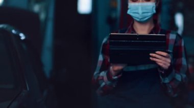 İş üniformalı, yüzünde tıbbi maske olan bir kadın tamircinin portresi tablette çalışıyor. Garajda..