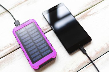 Modern teknoloji kavramı. Siyah ekranlı akıllı telefon, taşınabilir güneş enerjisi mor şarjlı. Çizgili beyaz ahşap arka plan. Kapat ve boşluğu kopyala.