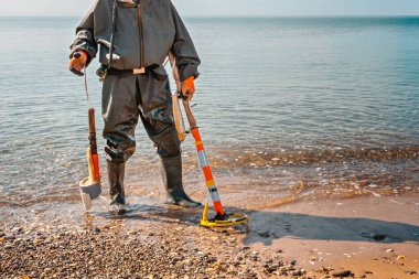 Metal dedektörlü bir adam plajda duruyor, yeni arkeolojik bulgulara hazır. Arka planda deniz.