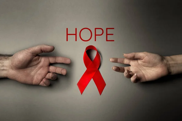 A female\'s hand and a male\'s hand reach for the red ribbon. Dark background. Flat lay. Inscription HOPE. The concept of world AIDS day.