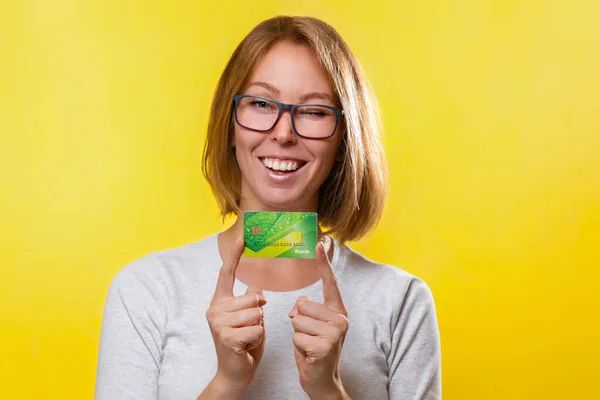 銀行カードと金融 眼鏡をかけた若いブロンドの女性が 両手の人差し指で銀行カードを持っている 黄色の背景 スペースのコピー — ストック写真