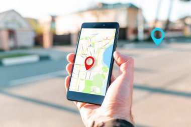 Erkek el, akıllı telefonu online kartla tutuyor ki bu da mekanın kırmızı bir simgesidir. Arka planda bulanık bir sokak ve mavi bir konum simgesi var. Çevrimiçi navigasyon ve GPS kavramı.