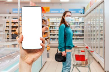 Ekranda alışveriş sepeti olan bir kişinin eli cep telefonunu tutuyor. Arka planda, bir süpermarkette bulanık bir şekilde tıbbi maske takan bir kadın var. Model yap. Online alışveriş kavramı.