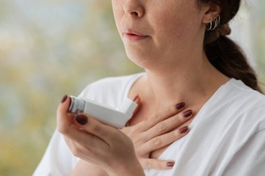 Allergic Asthma. A woman experiencing an asthmatic attack, holding an inhaler with medication to her face. Close up of hands. Blurry background. clipart