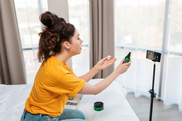 Social video. A beautiful female vlogger streaming live video on smartphone and showing a new cosmetic. Windows on the background. Side view.