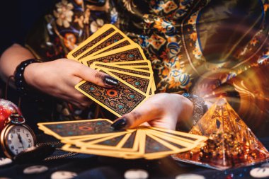 Astroloji ve ezoterizm. Bir falcının elinde Tarot kartları hayranı var. Masanın üzerinde rünler ve sihirli cam piramit var. Ellerin yakın çekimi.