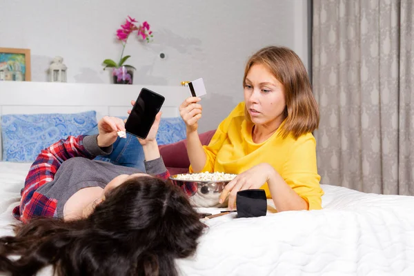 二人の若い女性がベッドの上に横たわっています スマートフォンを使用して 銀行カードを保持 オンラインショッピングと隔離の概念 — ストック写真