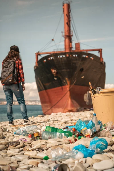 A woman with a backpack behind her back, standing on the background of the sea and the ship close-up. In the foreground is a dumpster and scattered plastic bottles. Vertical.