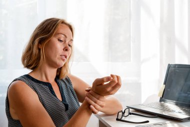 Masada oturan bir kadın bileklerini yoğuruyor. Arka planda beyaz pencere var. İş yerinde yorgunluk, karantina ve salgın sırasında kendini izole etme kavramı..