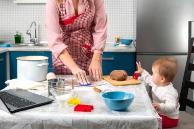 Önlüklü bir anne mutfakta kurabiye yapıyor. Bebek bir sandalyeye oturur ve süreci izler. Masada bir dizüstü bilgisayar var. Kapatın. Ev yemeği ve video iletişimi kavramı.