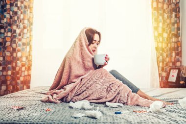 Hastalıklar ve mevsimsel soğuk algınlığı kavramı. Genç esmer bir kadın battaniyeye sarılı olarak yatakta oturuyor ve çay içiyor. Yanında mendiller, bir termometre ve haplar var. Arka plandaki güneş ışığı penceresi.