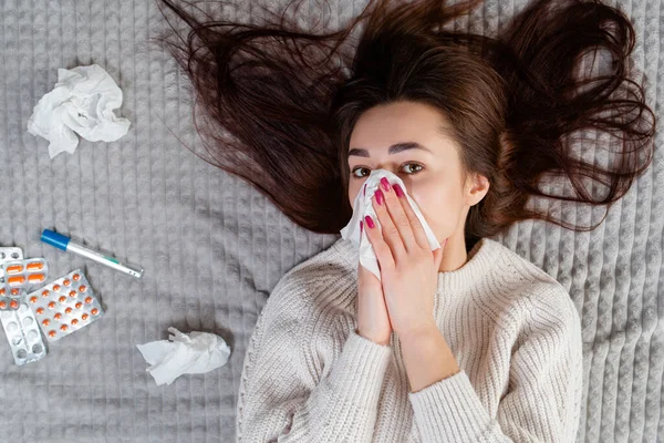 The concept of diseases and seasonal colds. A young brunette woman lies on the bed with her hair down and blows her nose. About not lie pills and thermometer. View from the top.