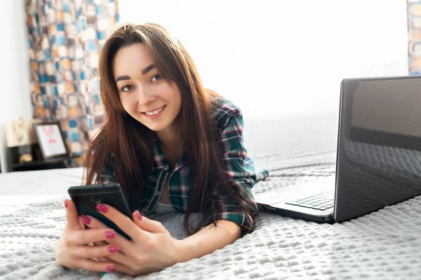 A young brunette woman with a joyful look lies on the bed and uses a smartphone, a laptop is lying next to her. In the background, the interior of the room and the window. Copy space.