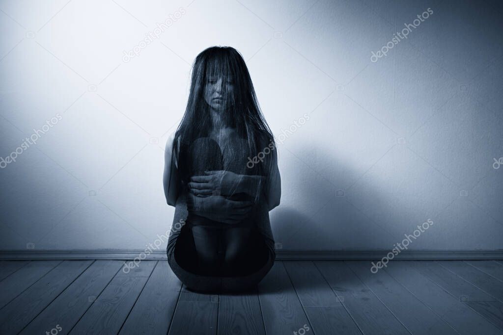 A young woman sits huddled on the floor of a room, covered by her hair. There is a light wall in the background.Human trafficking, sexual slavery concept. Copy space.