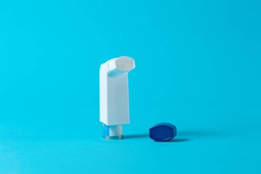 Astım tedavisinde kullanılıyor. Üzerinde ilaç olan açık bir astım spreyi mavi arka plandaki kapağın yanında duruyor..