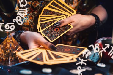 Astroloji ve kehanet. Bir falcının elinde Tarot kartları hayranı var. Köşelerde Zodiac işaretleri gösteriliyor. Masanın üzerinde rünler ve tılsımlar var. Ellerin yakın çekimi.