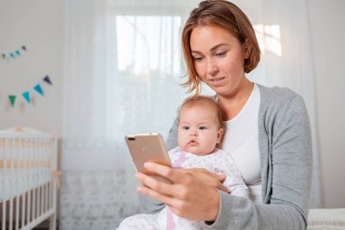 Genç annenin portresi kucağında sevimli bir bebek tutuyor ve akıllı telefonunda ona bir şey gösteriyor. Çevrimiçi eğlence kavramı.