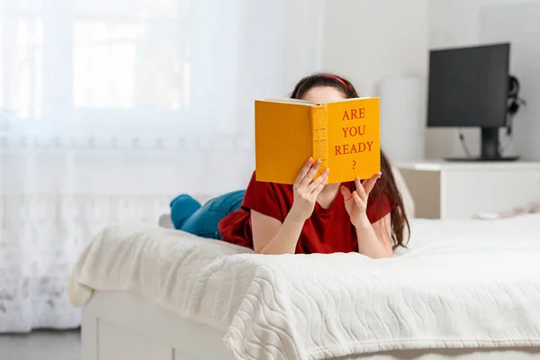 Młoda Kobieta Leży Łóżku Brzuchu Zakrywając Twarz Książką Napisem Jesteś — Zdjęcie stockowe