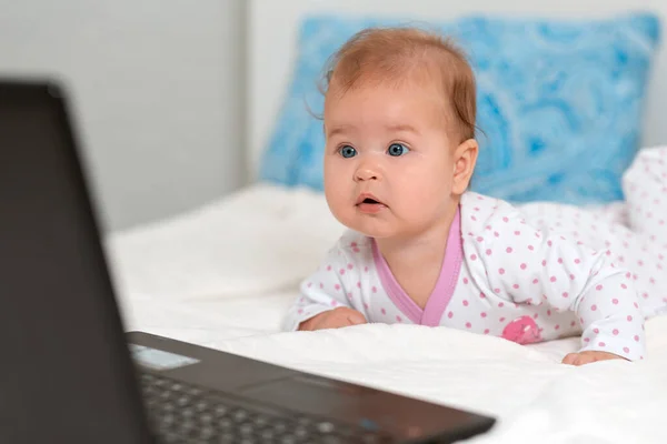 Portrait of amazement baby in sliders lies on the bed and looks at the laptop. The concept of teaching children to modern technologies.