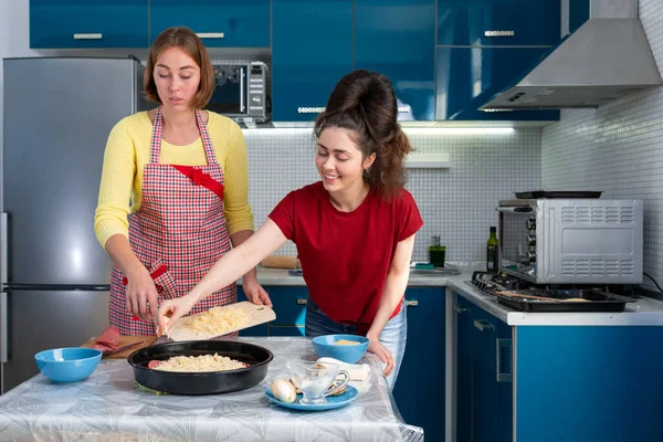 Two young women cook pizza, sprinkling cheese on top of it. Kitchen on the background. The concept of homemade food and lgbt couple.