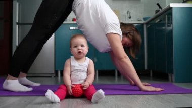 Çocuklu yoga. Genç bir anne yoga yapıyor ve bebek elinde bir oyuncakla minderin yanında oturuyor. Çocuklarla evde eğitim ve yoga kavramı.