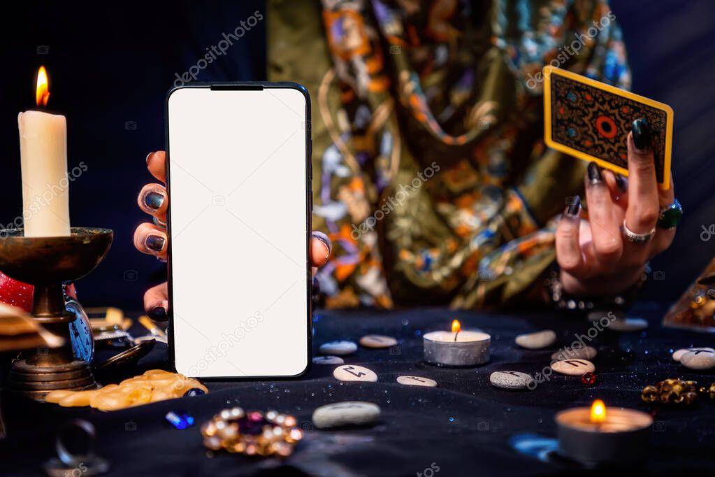 The fortune teller is holding a Tarot card and a smartphone with a white screen. Mock up. Close-up. The concept of divination, magic and esotericism.