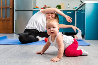 Çocuk yerde sürünüyordu. Arka planda, bulanık bir şekilde, genç bir anne mutfaktaki bir halıya uzanıyor. Çocuklarla evde spor eğitimi kavramı.