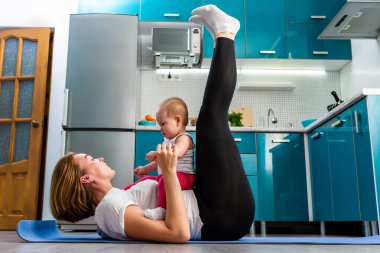 Bebekli bir anne, mutfakta paspasın üzerinde spor yaparken, bacaklarını kaldırırken eğleniyor. Çocuklarla evde spor eğitimi kavramı.