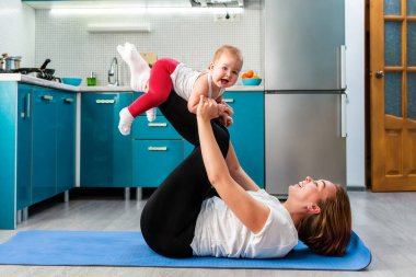 Gülümseyen bir anne, minderde spor yaparken sevimli mutlu bir bebeği kollarında tutar. Çocuklarla evde spor eğitimi kavramı.