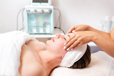 Kozmetik uzmanı bir kadına yüz ve kafa masajı prosedürü uygular. Arka planda kozmetik malzemeler var. Müşterinin portresi, yan görüş. Kozmetoloji ve tedavi kavramı.