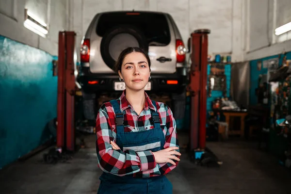 制服を着た若い女性整備士の肖像画が腕を組んでポーズをとった 後ろには自動修理工場とリフトの上の車があります — ストック写真